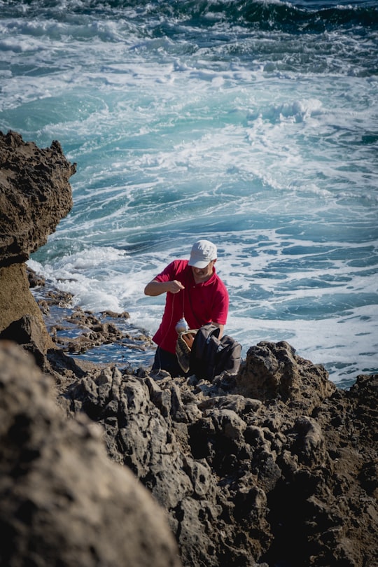 man in red jacket sitting on rock formation near sea during daytime in Boca do Inferno Portugal