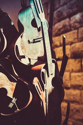Several vintage, rustic guitars are stacked together, displaying their intricate curves and wooden textures. The lighting casts warm, dramatic shadows on the wall behind, highlighting the contrast between the instruments and the stone surface.