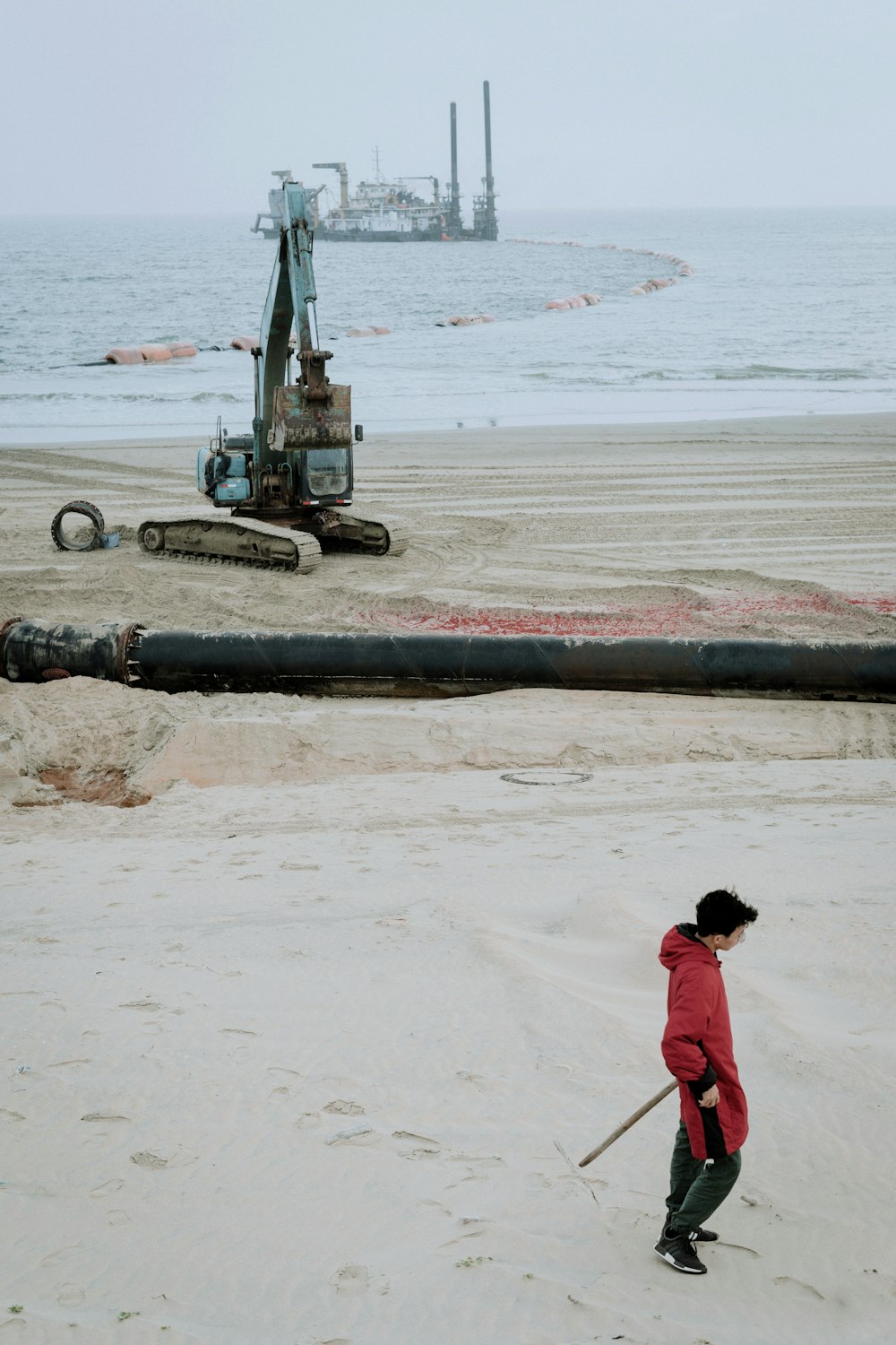 woman in red hoodie standing on beach near black heavy equipment during daytime