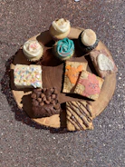 An assortment of cupcakes and baked goods are displayed on a round wooden board. The cupcakes feature intricate frosting designs in white and turquoise, while the baked items below include decorated pastries, a chocolate brownie topped with chocolate chips, and a cookie with chocolate drizzle.