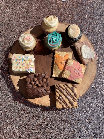 An assortment of cupcakes and baked goods are displayed on a round wooden board. The cupcakes feature intricate frosting designs in white and turquoise, while the baked items below include decorated pastries, a chocolate brownie topped with chocolate chips, and a cookie with chocolate drizzle.