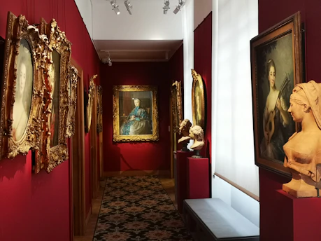 An art gallery hallway with richly decorated walls painted in deep red. Several framed paintings and portraits in ornate gold frames adorn the walls. On pedestals, there are classical-style busts. The lighting is focused on the artworks, creating an intimate atmosphere.