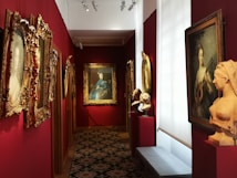 An art gallery hallway with richly decorated walls painted in deep red. Several framed paintings and portraits in ornate gold frames adorn the walls. On pedestals, there are classical-style busts. The lighting is focused on the artworks, creating an intimate atmosphere.