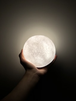 A hand is gently holding a glowing sphere that resembles the moon, with intricate crater-like textures. The background is dark, emphasizing the soft light emitted from the spherical object.