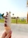 A hand holds a soft-serve ice cream cone with swirls of chocolate and vanilla in front of a grassy park area. Palm trees and people sitting on the grass can be seen in the background, with a glimpse of a body of water under a clear sky.