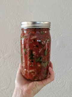 A hand is holding a clear glass jar filled with a chunky, red salsa. The salsa contains visible pieces of tomatoes, herbs, and possibly onions. The jar has a metal screw-on lid.