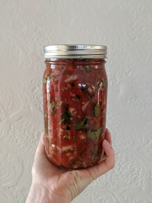 A hand is holding a clear glass jar filled with a chunky, red salsa. The salsa contains visible pieces of tomatoes, herbs, and possibly onions. The jar has a metal screw-on lid.