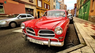 A classic red Volvo car is parked on a street lined with colorful buildings. The car's chrome details and vintage design stand out. The street is narrow with a mix of other parked cars, and the sky appears overcast.