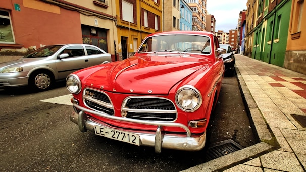 A classic red Volvo car is parked on a street lined with colorful buildings. The car's chrome details and vintage design stand out. The street is narrow with a mix of other parked cars, and the sky appears overcast.