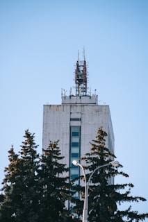 A tall, modern building is equipped with numerous antennas and communication equipment on its roof. In the foreground, several evergreen trees partially obscure the lower portion of the building, while a lamp post is visible among the trees. The sky is clear and blue, providing a stark contrast to the concrete structure.