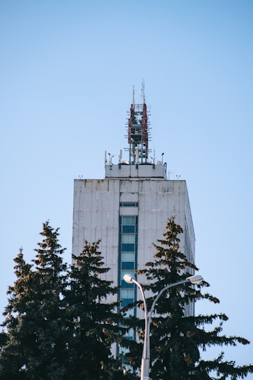 A tall, modern building is equipped with numerous antennas and communication equipment on its roof. In the foreground, several evergreen trees partially obscure the lower portion of the building, while a lamp post is visible among the trees. The sky is clear and blue, providing a stark contrast to the concrete structure.