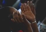 Close-up of hands raised in prayer with sunlight streaming through stained glass windows.