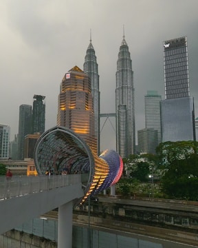 A cityscape featuring two tall, identical skyscrapers with a unique twisted bridge or walkway in the foreground. The structures are illuminated with warm lighting against a cloudy sky.