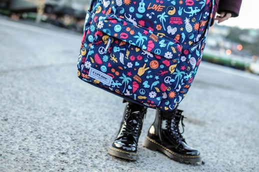 A colorful backpack adorned with various playful and vivid designs, such as peace symbols, palm trees, flowers, and other quirky icons. The backpack is worn by a person standing on a concrete surface, visible from the knees down, wearing shiny black boots.