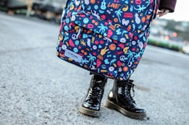 A colorful backpack adorned with various playful and vivid designs, such as peace symbols, palm trees, flowers, and other quirky icons. The backpack is worn by a person standing on a concrete surface, visible from the knees down, wearing shiny black boots.