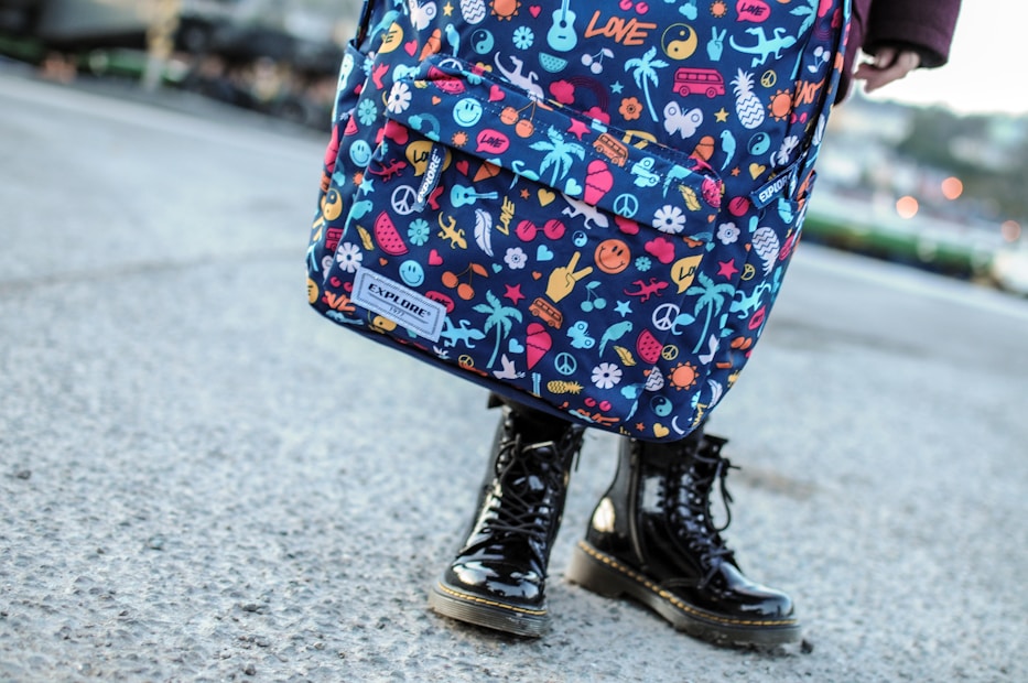 A colorful backpack adorned with various playful and vivid designs, such as peace symbols, palm trees, flowers, and other quirky icons. The backpack is worn by a person standing on a concrete surface, visible from the knees down, wearing shiny black boots.