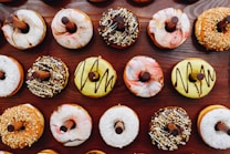 A variety of donuts are arranged in rows on a wooden surface. The donuts have different toppings, including chocolate drizzle, nuts, coconut flakes, and colorful glazes. Each donut has a stick inserted in the center.