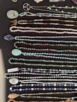 A collection of various beaded necklaces displayed on a dark surface. The necklaces feature a range of bead sizes and colors, including turquoise, gold, and earthy tones. Some necklaces have unique pendants and charms alongside the beads.