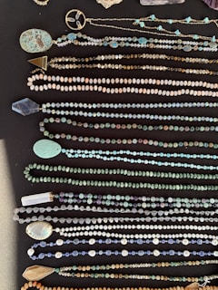 A collection of various beaded necklaces displayed on a dark surface. The necklaces feature a range of bead sizes and colors, including turquoise, gold, and earthy tones. Some necklaces have unique pendants and charms alongside the beads.