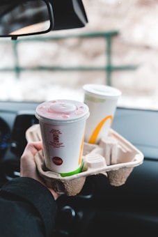 A person is holding a cardboard cup holder with two large disposable drink cups inside a vehicle. The cups have plastic lids and are likely from a fast-food restaurant. The background includes the car's interior, rearview mirror, and a blurry exterior view through the window.