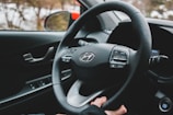 The interior of a car is shown, featuring a steering wheel with the Hyundai logo in the center. Several control buttons are visible on the steering wheel, and a driver's hand is lightly holding it. The car's dashboard includes visible elements such as an engine start/stop button and a side mirror.