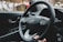 The interior of a car is shown, featuring a steering wheel with the Hyundai logo in the center. Several control buttons are visible on the steering wheel, and a driver's hand is lightly holding it. The car's dashboard includes visible elements such as an engine start/stop button and a side mirror.