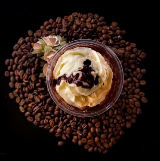 A top view of a dessert cup filled with whipped cream and drizzled with chocolate sauce, surrounded by a circle of coffee beans and a small decorative flower.