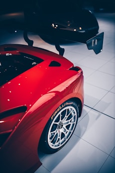 A close-up view of a red sports car featuring a prominent rear wing and large Pirelli-branded tires on silver alloy wheels. The car is positioned on a glossy white tiled floor with reflections visible.