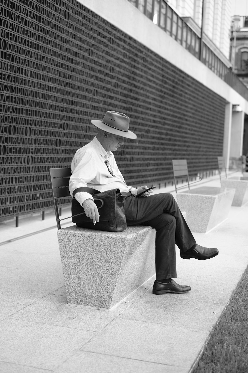 grayscale photo of man in white long sleeve shirt and black pants sitting on concrete bench