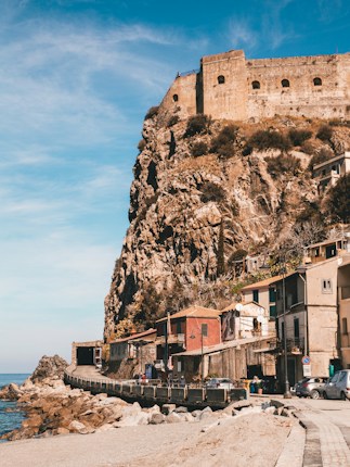 A rustic coastal village with colorful houses nestled against a large rocky cliff. Atop the cliff stands an ancient stone fortress with small windows, providing a striking contrast with the clear blue sky. The foreground shows a narrow road lined with a few parked cars and some pedestrians.