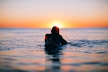 Sunset silhouette of a surfer sitting on their board, watching the ocean.