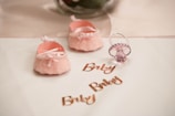 Two soft pink baby shoes with bows and pearl decorations are placed on a white surface next to a pink pacifier. The word 'Baby' is written multiple times in shiny rose gold lettering.