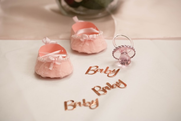 Two soft pink baby shoes with bows and pearl decorations are placed on a white surface next to a pink pacifier. The word 'Baby' is written multiple times in shiny rose gold lettering.