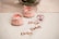 Two soft pink baby shoes with bows and pearl decorations are placed on a white surface next to a pink pacifier. The word 'Baby' is written multiple times in shiny rose gold lettering.