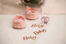 Two soft pink baby shoes with bows and pearl decorations are placed on a white surface next to a pink pacifier. The word 'Baby' is written multiple times in shiny rose gold lettering.