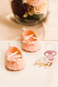 A pair of delicate pink baby shoes with lacy bows and pearl embellishments are placed on a soft surface next to a pink pacifier adorned with rhinestones. Behind them, a glass jar contains a bouquet of artificial roses in various colors, including deep red, pastel pink, and cream, with green leaves.