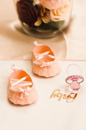 A pair of delicate pink baby shoes with lacy bows and pearl embellishments are placed on a soft surface next to a pink pacifier adorned with rhinestones. Behind them, a glass jar contains a bouquet of artificial roses in various colors, including deep red, pastel pink, and cream, with green leaves.
