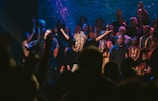 A group of people are gathered on a stage, some raising their hands and others singing or speaking into microphones. The lighting creates a vibrant atmosphere with blue and purple hues, and everyone appears to be participating in a shared event or performance.