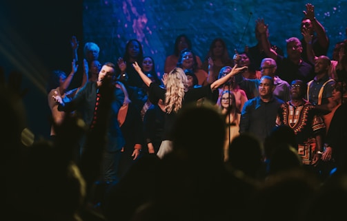 A group of people are gathered on a stage, some raising their hands and others singing or speaking into microphones. The lighting creates a vibrant atmosphere with blue and purple hues, and everyone appears to be participating in a shared event or performance.