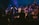 A group of people are gathered on a stage, some raising their hands and others singing or speaking into microphones. The lighting creates a vibrant atmosphere with blue and purple hues, and everyone appears to be participating in a shared event or performance.