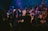 A group of people are gathered on a stage, some raising their hands and others singing or speaking into microphones. The lighting creates a vibrant atmosphere with blue and purple hues, and everyone appears to be participating in a shared event or performance.