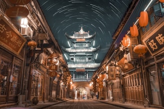 An ancient Asian street scene under a night sky filled with star trails. Traditional architecture is lit up, with a large, ornate tower in the background. The street is lined with wooden buildings adorned with glowing red lanterns and various signage. The atmosphere is serene and mystical.