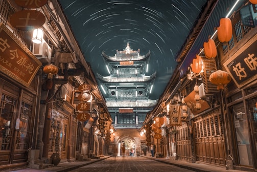 An ancient Asian street scene under a night sky filled with star trails. Traditional architecture is lit up, with a large, ornate tower in the background. The street is lined with wooden buildings adorned with glowing red lanterns and various signage. The atmosphere is serene and mystical.