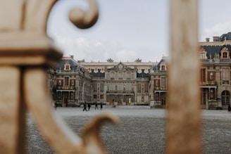 An ornate, historical palace with intricate architectural details, seen through the bars of a golden gate. The cobblestone courtyard is mostly empty, with a few people walking in the distance under an overcast sky. The building features classic European design with decorative elements in gold, brick, and stone.
