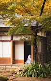 A traditional Japanese house surrounded by lush greenery, with a person dressed in a white garment sitting inside, visible through an open doorway.