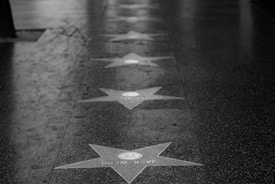 A black and white image of a sidewalk featuring Hollywood Walk of Fame stars. The stars are arranged in a line, with names inscribed on them. The surface appears wet, suggesting recent rain or weather exposure.