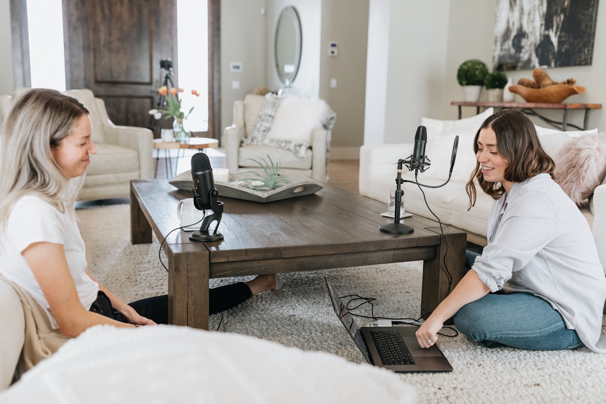 Two podcasters sitting with mics, on a rug in a living room, across a low-rise table. One of them is operating a laptop. 