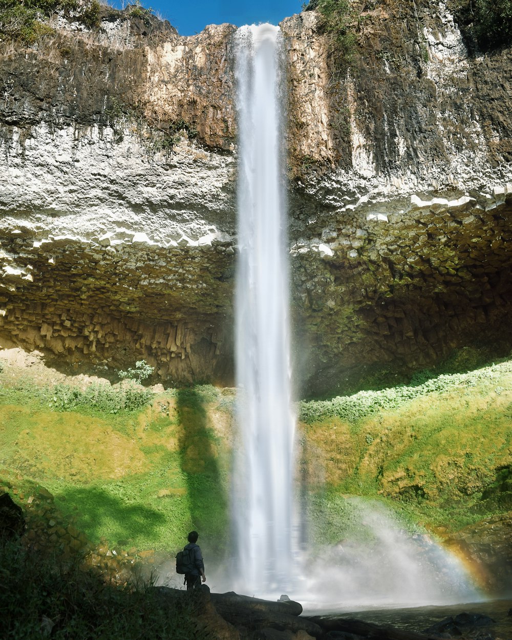 person in black jacket standing on green grass field near waterfalls during daytime