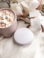 A cozy scene showing MGGSkin moisturizer and cleanser on a pastel pink towel beside a cup of tea.