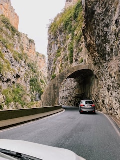 A narrow winding road passes through a rocky canyon with towering cliffs on either side. The road curves around a stone archway that is part of the canyon wall. A dark-colored car is driving ahead, navigating the scenic route surrounded by rugged natural beauty. The cliffs are covered in patches of green vegetation.